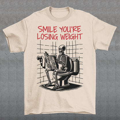 A humorous t-shirt design featuring a cartoon skeleton sitting on a toilet, legs crossed, casually reading a newspaper. The skeleton has a relaxed, almost cheerful expression, emphasizing the absurdity of the scene. Above the image, bold text reads: "SMILE YOU'RE LOSING WEIGHT," playing on the double meaning of weight loss and the skeleton’s bony frame. The design blends bathroom humor with ironic positivity for a lighthearted laugh.