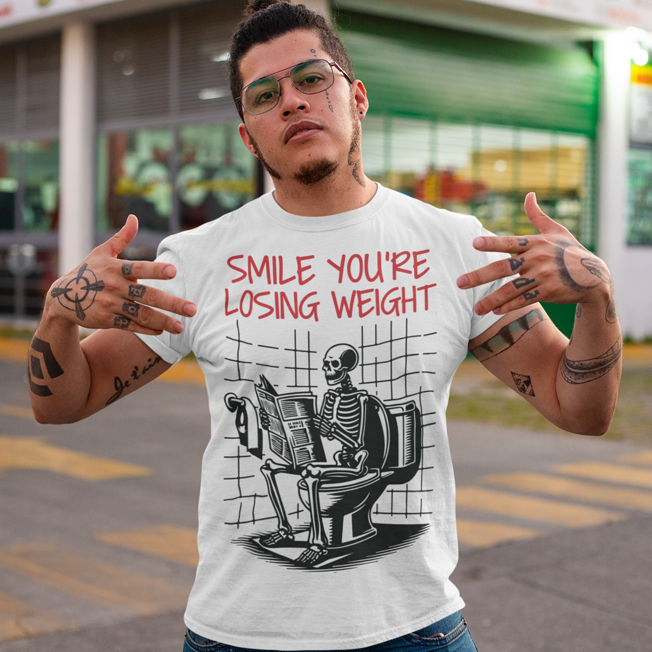 A humorous t-shirt design featuring a cartoon skeleton sitting on a toilet, legs crossed, casually reading a newspaper. The skeleton has a relaxed, almost cheerful expression, emphasizing the absurdity of the scene. Above the image, bold text reads: "SMILE YOU'RE LOSING WEIGHT," playing on the double meaning of weight loss and the skeleton’s bony frame. The design blends bathroom humor with ironic positivity for a lighthearted laugh.
