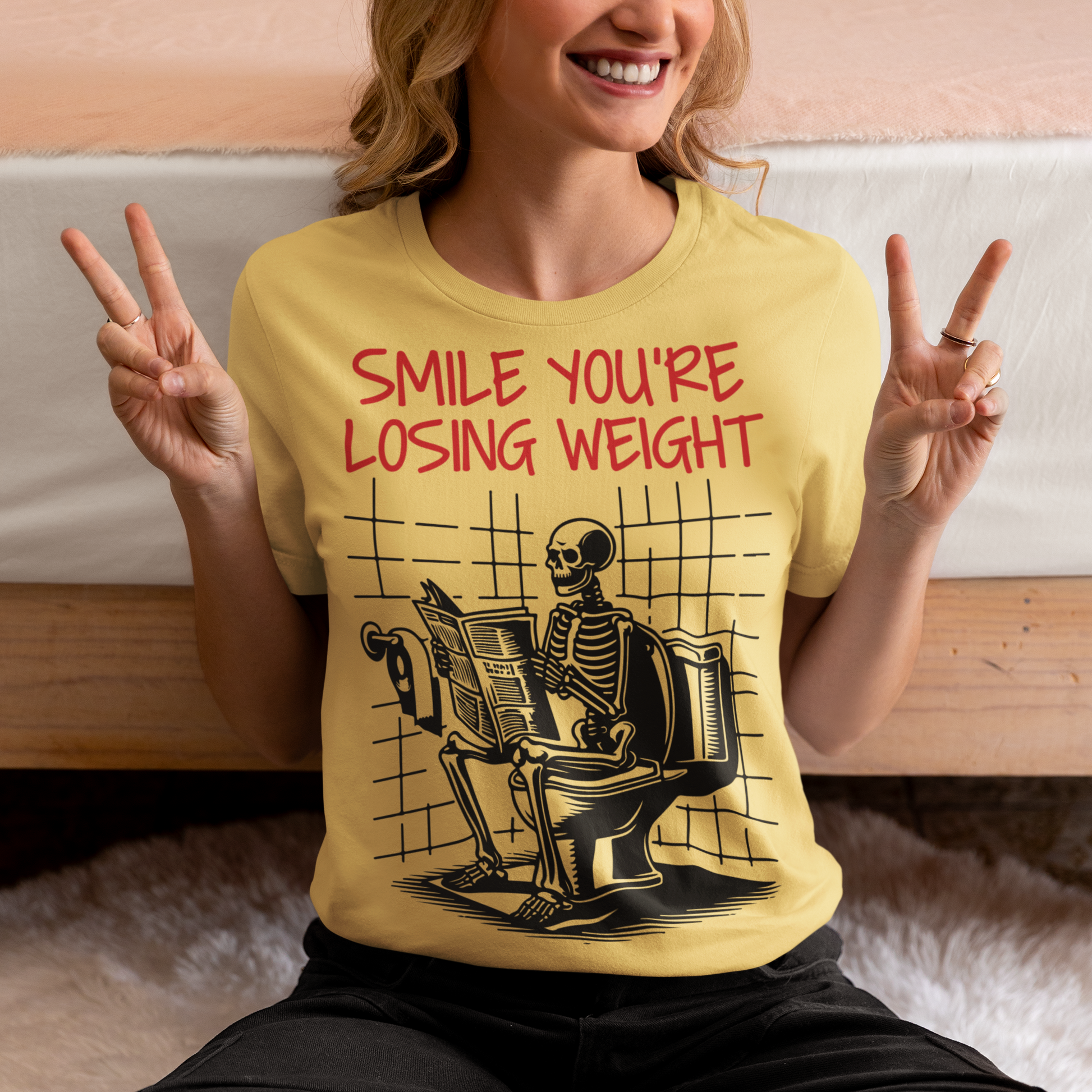 A humorous t-shirt design featuring a cartoon skeleton sitting on a toilet, legs crossed, casually reading a newspaper. The skeleton has a relaxed, almost cheerful expression, emphasizing the absurdity of the scene. Above the image, bold text reads: "SMILE YOU'RE LOSING WEIGHT," playing on the double meaning of weight loss and the skeleton’s bony frame. The design blends bathroom humor with ironic positivity for a lighthearted laugh.
