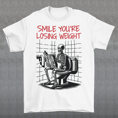 A humorous t-shirt design featuring a cartoon skeleton sitting on a toilet, legs crossed, casually reading a newspaper. The skeleton has a relaxed, almost cheerful expression, emphasizing the absurdity of the scene. Above the image, bold text reads: "SMILE YOU'RE LOSING WEIGHT," playing on the double meaning of weight loss and the skeleton’s bony frame. The design blends bathroom humor with ironic positivity for a lighthearted laugh.