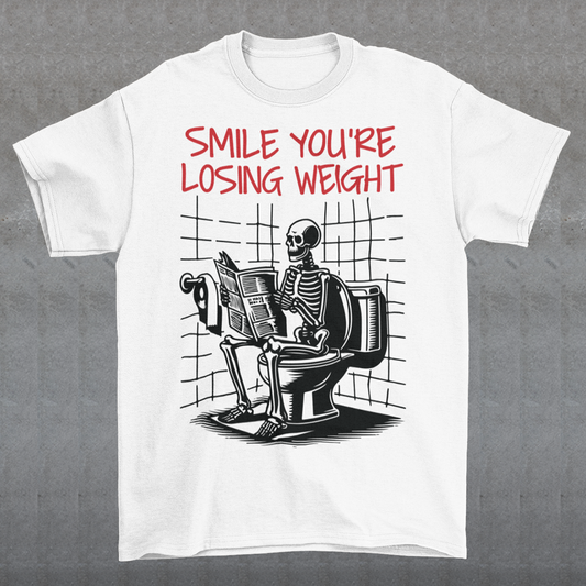 A humorous t-shirt design featuring a cartoon skeleton sitting on a toilet, legs crossed, casually reading a newspaper. The skeleton has a relaxed, almost cheerful expression, emphasizing the absurdity of the scene. Above the image, bold text reads: "SMILE YOU'RE LOSING WEIGHT," playing on the double meaning of weight loss and the skeleton’s bony frame. The design blends bathroom humor with ironic positivity for a lighthearted laugh.