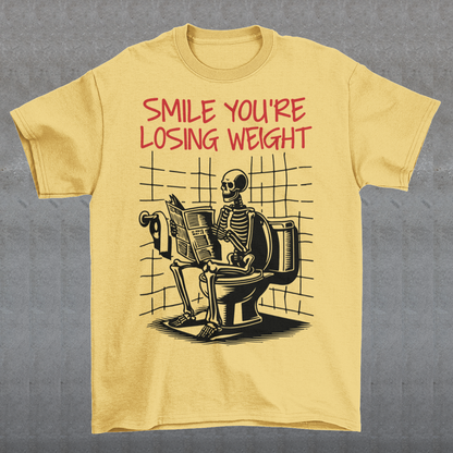 A humorous t-shirt design featuring a cartoon skeleton sitting on a toilet, legs crossed, casually reading a newspaper. The skeleton has a relaxed, almost cheerful expression, emphasizing the absurdity of the scene. Above the image, bold text reads: "SMILE YOU'RE LOSING WEIGHT," playing on the double meaning of weight loss and the skeleton’s bony frame. The design blends bathroom humor with ironic positivity for a lighthearted laugh.