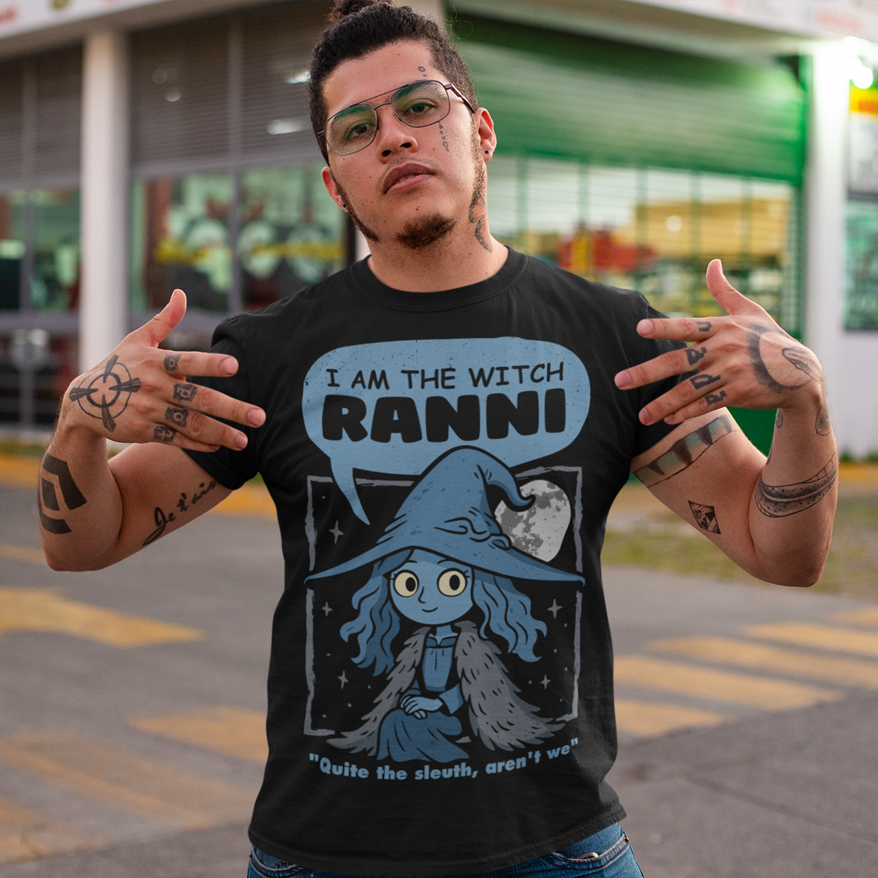 Black t-shirt featuring a cute blue witch character with large eyes wearing a pointed hat and cloak, set against a night sky with a full moon and stars. Text above reads “I AM THE WITCH RANNI,” with a smaller quote below saying “Quite the sleuth, aren’t we.” The design has a dark fantasy, game-inspired style with soft blue tones and a whimsical, mystical aesthetic.