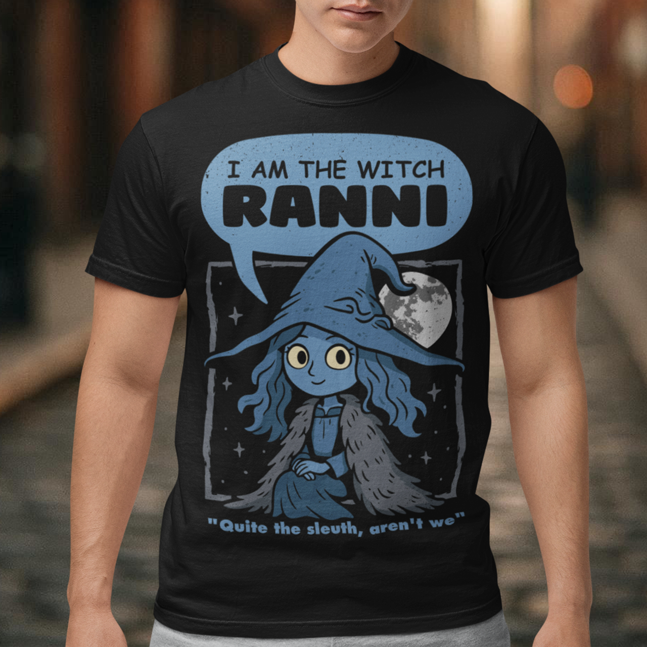 Black t-shirt featuring a cute blue witch character with large eyes wearing a pointed hat and cloak, set against a night sky with a full moon and stars. Text above reads “I AM THE WITCH RANNI,” with a smaller quote below saying “Quite the sleuth, aren’t we.” The design has a dark fantasy, game-inspired style with soft blue tones and a whimsical, mystical aesthetic.