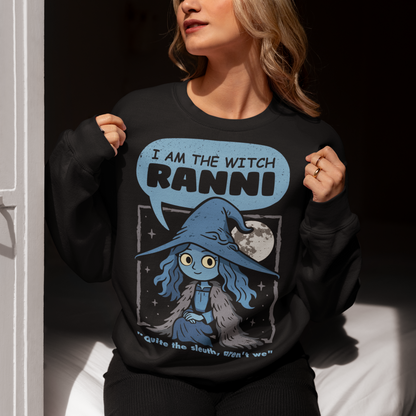 Black sweatshirt featuring a cute blue witch character with large eyes wearing a pointed hat and cloak, set against a night sky with a full moon and stars. Text above reads “I AM THE WITCH RANNI,” with a smaller quote below saying “Quite the sleuth, aren’t we.” The design has a dark fantasy, game-inspired style with soft blue tones and a whimsical, mystical aesthetic.