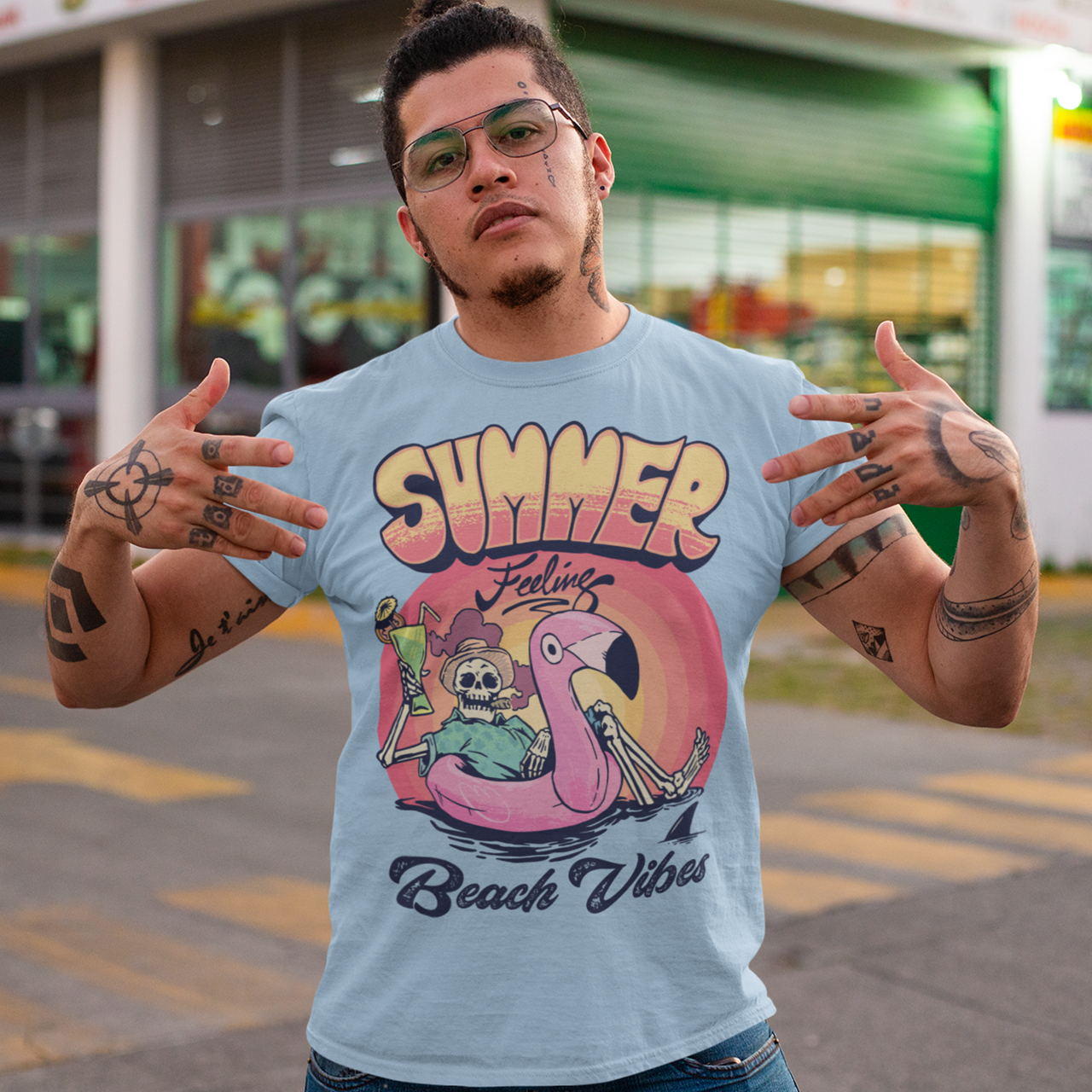 Light blue t-shirt of a skeleton floating on a rubber flamingo in the sea with a cocktail and cigar in front of a sunset with text "SUMMER FEELING BEACH VIBES".