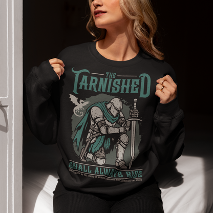 Black sweatshirt featuring a dark fantasy design of a kneeling armored knight holding a sword with teal accents and a dragon silhouette in the background. Large text reads “The Tarnished Shall Always Rise,” with smaller medieval-style text underneath. Inspired by fantasy RPG and soulslike gaming aesthetics.