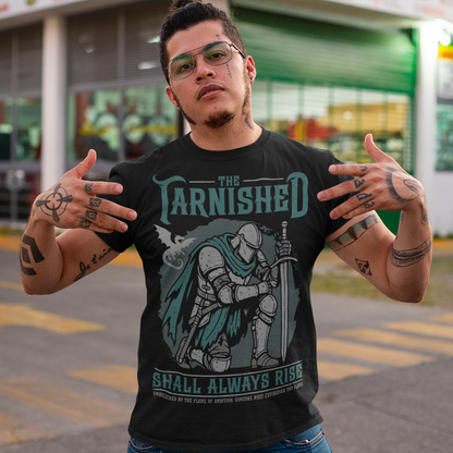 Black t-shirt featuring a dark fantasy design of a kneeling armored knight holding a sword with teal accents and a dragon silhouette in the background. Large text reads “The Tarnished Shall Always Rise,” with smaller medieval-style text underneath. Inspired by fantasy RPG and soulslike gaming aesthetics.