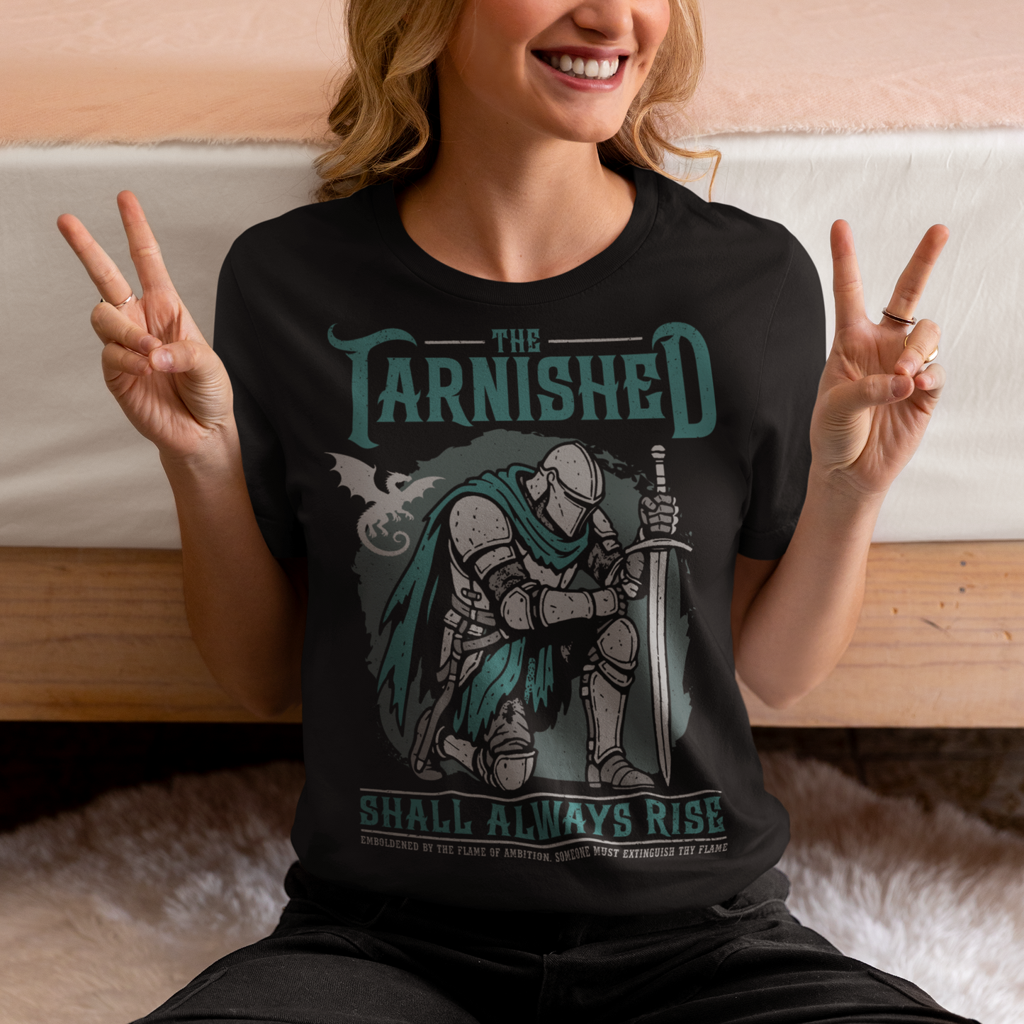 Black t-shirt featuring a dark fantasy design of a kneeling armored knight holding a sword with teal accents and a dragon silhouette in the background. Large text reads “The Tarnished Shall Always Rise,” with smaller medieval-style text underneath. Inspired by fantasy RPG and soulslike gaming aesthetics.
