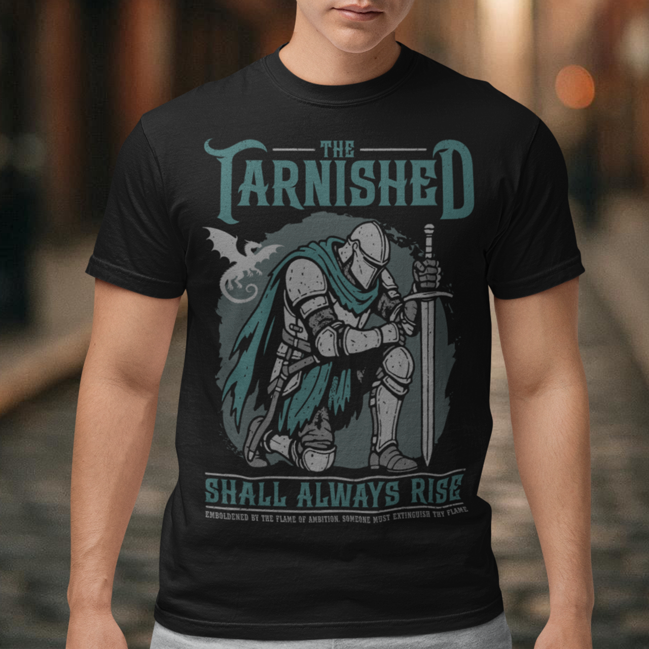 Black t-shirt featuring a dark fantasy design of a kneeling armored knight holding a sword with teal accents and a dragon silhouette in the background. Large text reads “The Tarnished Shall Always Rise,” with smaller medieval-style text underneath. Inspired by fantasy RPG and soulslike gaming aesthetics.