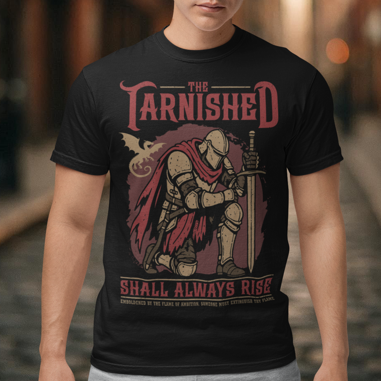 Black t-shirt featuring a dark fantasy design of a kneeling armored knight holding a sword with red accents and a dragon silhouette in the background. Large text reads “The Tarnished Shall Always Rise,” with smaller medieval-style text underneath. Inspired by fantasy RPG and soulslike gaming aesthetics.