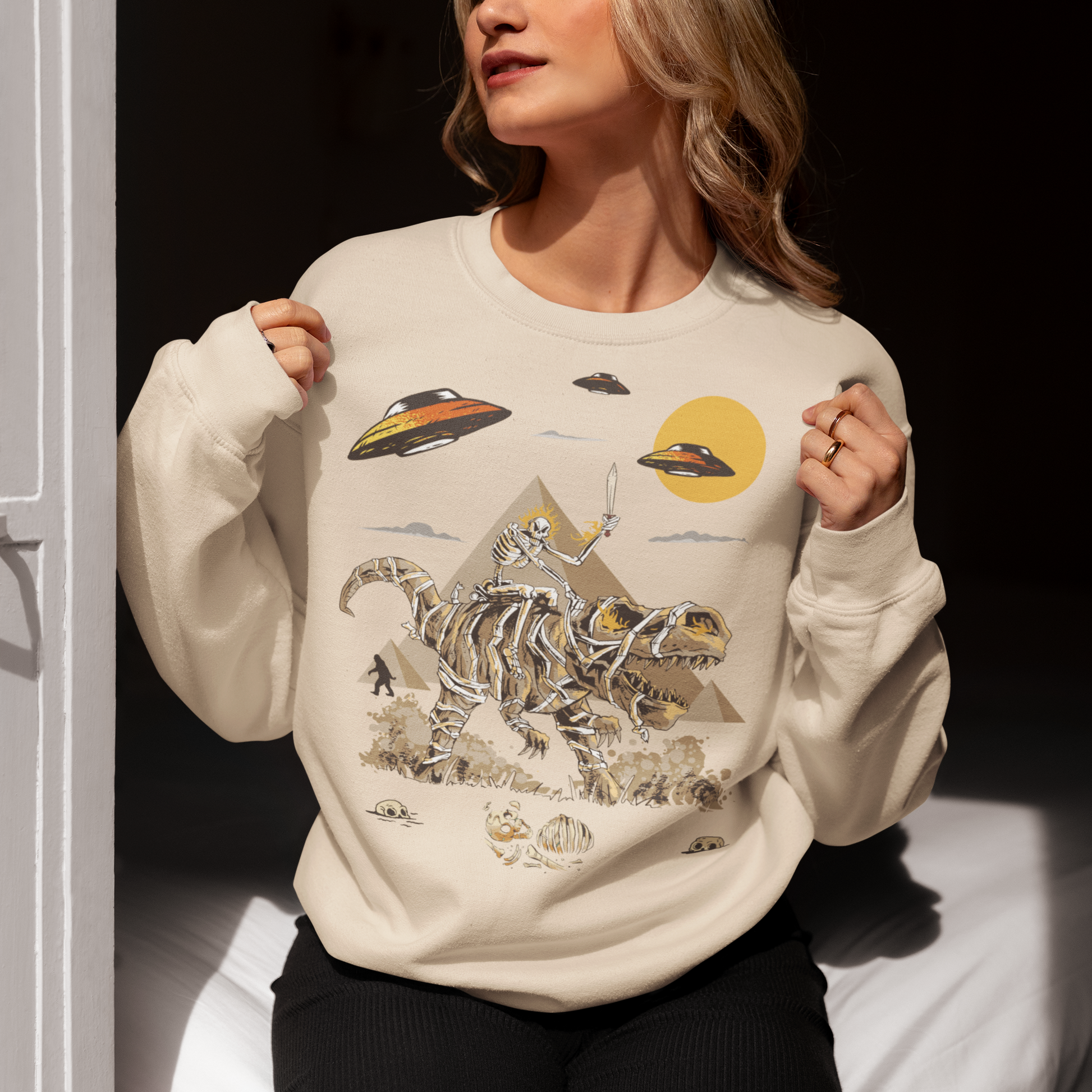 Cream sweatshirt with a skeleton riding a mummy dinosaur in ancient Egypt with UFO and bigfoot walking in the background.