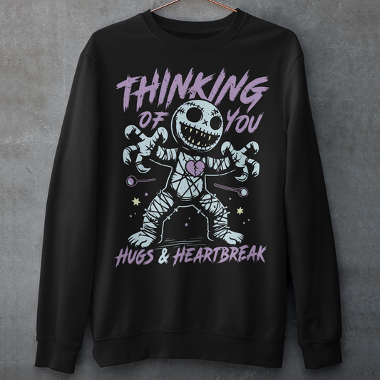 Dark humor meets twisted sweetness with this bold sweatshirt. Featuring a stitched-up voodoo doll at the center, the design captures that perfect mix of love and pain. Above the doll, the phrase “THINKING OF YOU” adds a playful sting, while below, “HUGS &amp; HEARTBREAK” ties it all together with equal parts irony and affection. Perfect for anyone who loves edgy, alternative fashion—or just wants to send a cheeky message without saying a word.