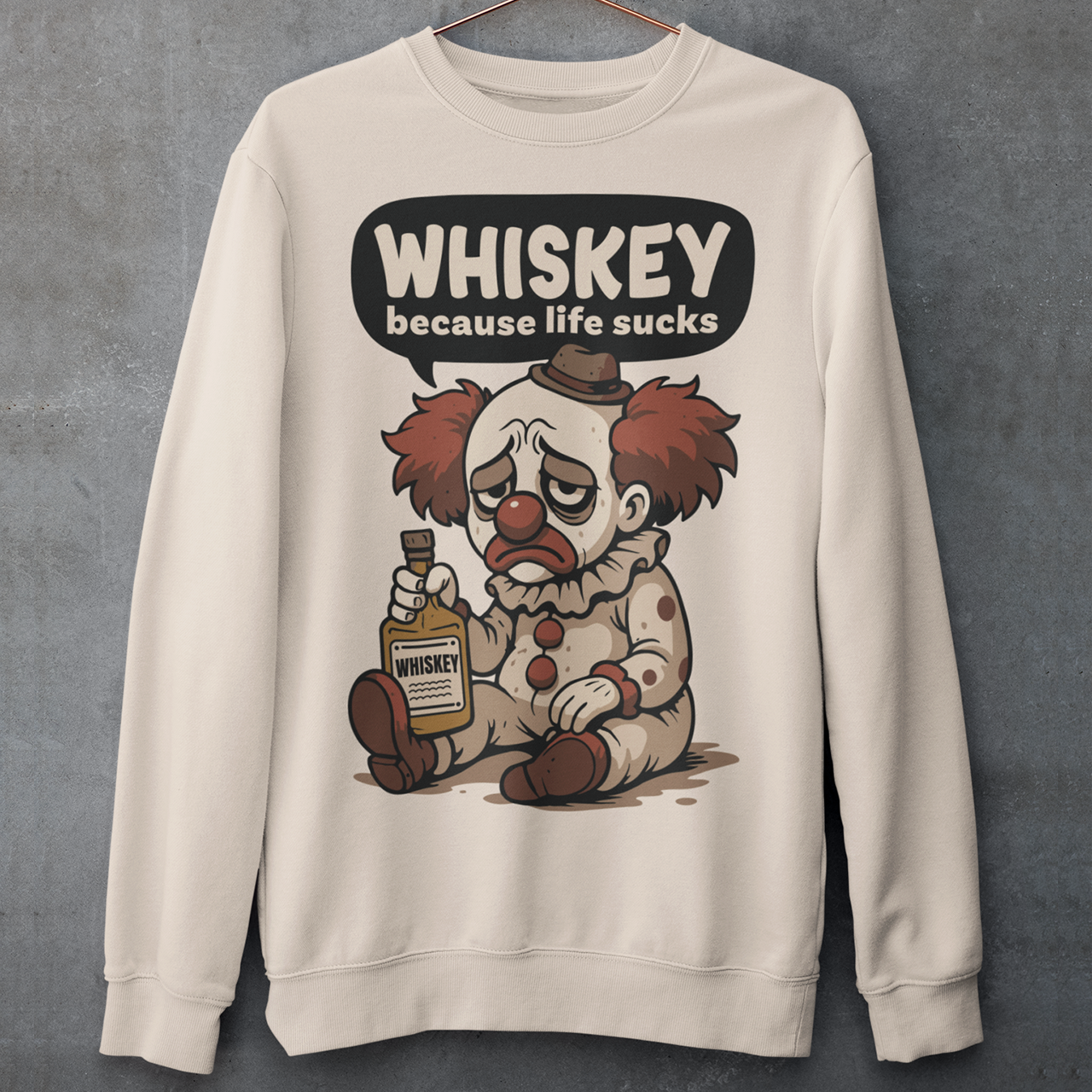 Beige sweatshirt featuring a sad cartoon clown with messy red hair and a tired expression, sitting on the ground holding a bottle labeled whiskey. Above the character, bold text reads “Whiskey because life sucks.” The design combines dark humor with a playful cartoon style, creating a sarcastic and slightly edgy visual.