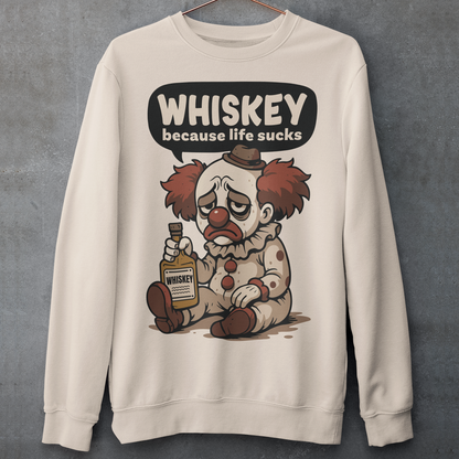 Beige sweatshirt featuring a sad cartoon clown with messy red hair and a tired expression, sitting on the ground holding a bottle labeled whiskey. Above the character, bold text reads “Whiskey because life sucks.” The design combines dark humor with a playful cartoon style, creating a sarcastic and slightly edgy visual.