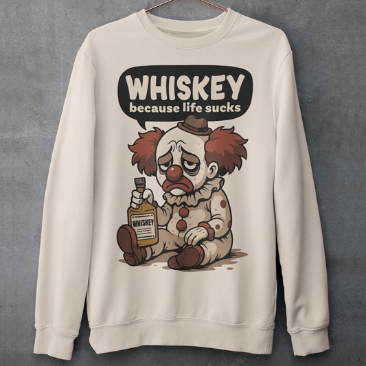 Beige sweatshirt featuring a sad cartoon clown with messy red hair and a tired expression, sitting on the ground holding a bottle labeled whiskey. Above the character, bold text reads “Whiskey because life sucks.” The design combines dark humor with a playful cartoon style, creating a sarcastic and slightly edgy visual.