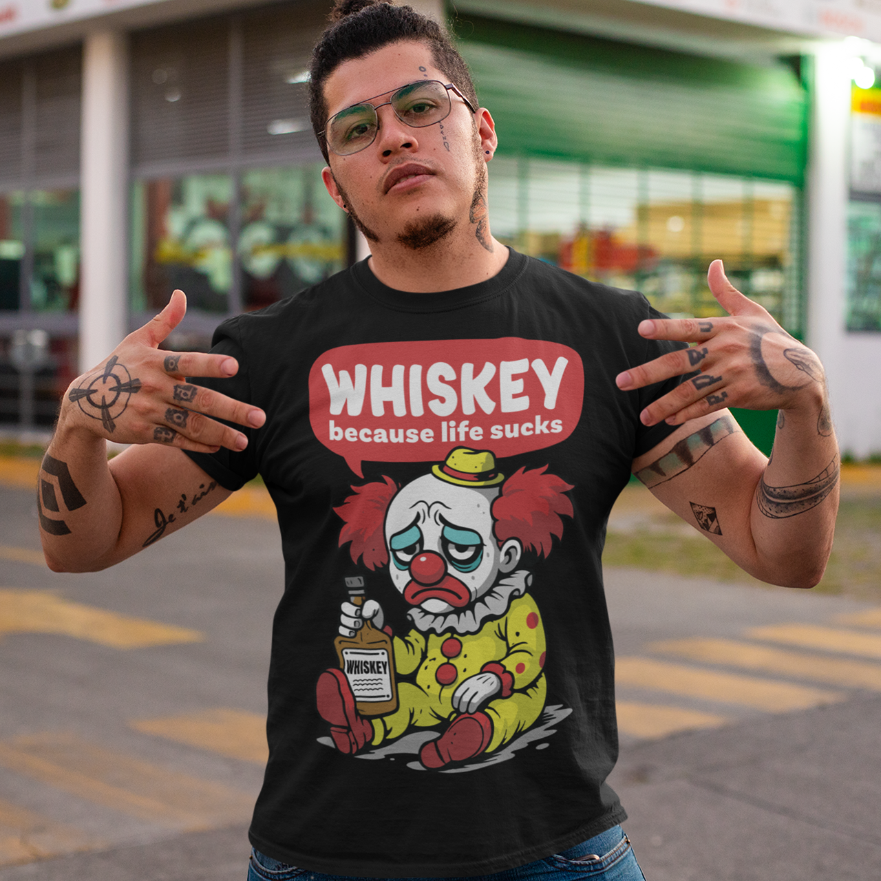 Black t-shirt featuring a sad cartoon clown with messy red hair and a tired expression, sitting on the ground holding a bottle labeled whiskey. Above the character, bold text reads “Whiskey because life sucks.” The design combines dark humor with a playful cartoon style, creating a sarcastic and slightly edgy visual.