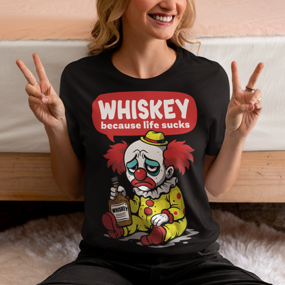 Black t-shirt featuring a sad cartoon clown with messy red hair and a tired expression, sitting on the ground holding a bottle labeled whiskey. Above the character, bold text reads “Whiskey because life sucks.” The design combines dark humor with a playful cartoon style, creating a sarcastic and slightly edgy visual.