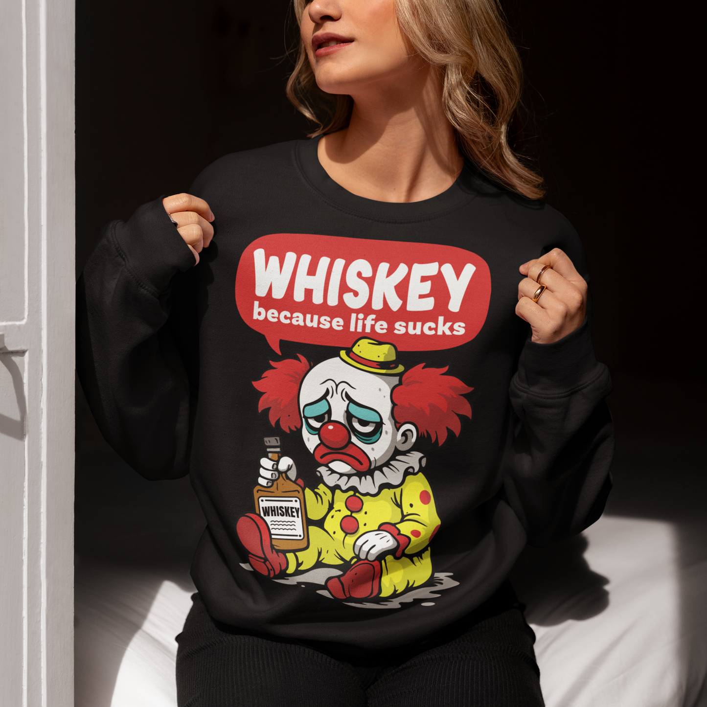 Black sweatshirt featuring a sad cartoon clown with messy red hair and a tired expression, sitting on the ground holding a bottle labeled whiskey. Above the character, bold text reads “Whiskey because life sucks.” The design combines dark humor with a playful cartoon style, creating a sarcastic and slightly edgy visual.
