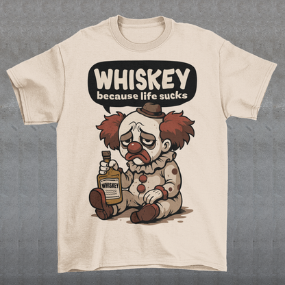 Cream t-shirt featuring a sad cartoon clown with messy red hair and a tired expression, sitting on the ground holding a bottle labeled whiskey. Above the character, bold text reads “Whiskey because life sucks.” The design combines dark humor with a playful cartoon style, creating a sarcastic and slightly edgy visual.