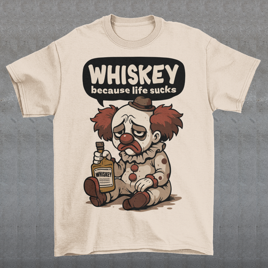 Cream t-shirt featuring a sad cartoon clown with messy red hair and a tired expression, sitting on the ground holding a bottle labeled whiskey. Above the character, bold text reads “Whiskey because life sucks.” The design combines dark humor with a playful cartoon style, creating a sarcastic and slightly edgy visual.