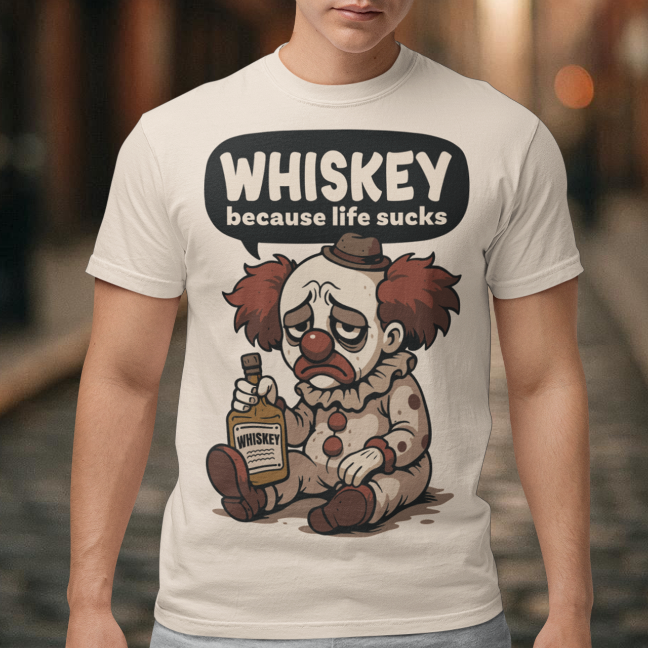 Cream t-shirt featuring a sad cartoon clown with messy red hair and a tired expression, sitting on the ground holding a bottle labeled whiskey. Above the character, bold text reads “Whiskey because life sucks.” The design combines dark humor with a playful cartoon style, creating a sarcastic and slightly edgy visual.