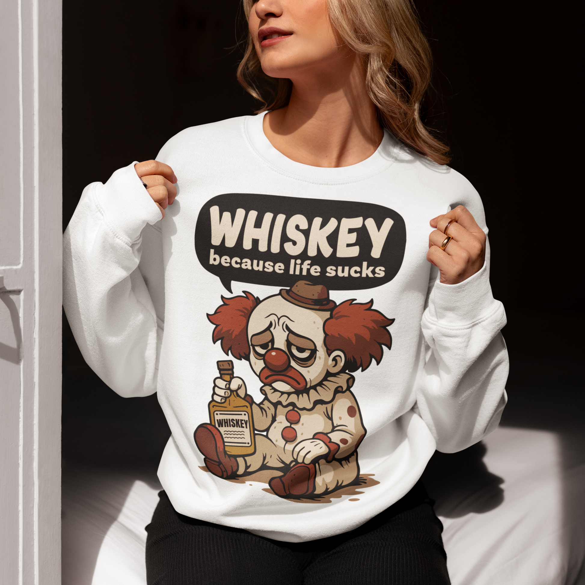 White sweatshirt featuring a sad cartoon clown with messy red hair and a tired expression, sitting on the ground holding a bottle labeled whiskey. Above the character, bold text reads “Whiskey because life sucks.” The design combines dark humor with a playful cartoon style, creating a sarcastic and slightly edgy visual.